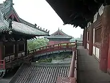 Several wooden buildings with grey tile roofs, connected by an arched bridge. A forest can be seen behind the buildings.