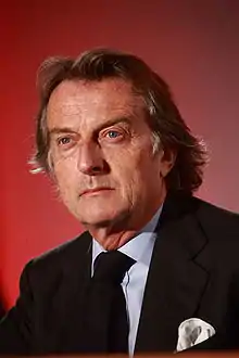 Upper body of a man in his sixties with neatly combed, brown but greying hair, wearing a smart grey suit and navy tie with small white spots. He is sitting at a table, upon which his hands rest.