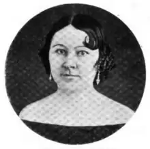 Portrait photograph of a middle-aged woman with her dark hair in an up-do, who is wearing drop earrings and an off-the-shoulder dark blouse.