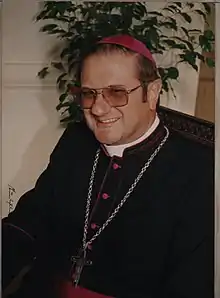 Eichhorn with eyeglasses is seated indoors facing left and smiling. He is wearing a bishop's black cassock with amaranth red piping and a zucchetto (also in amaranth red) as well as a pectoral cross suspended by a chain around his neck.