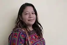 A woman with long brown hair wearing an embroidered top in three-quarter view smiles at the camera.