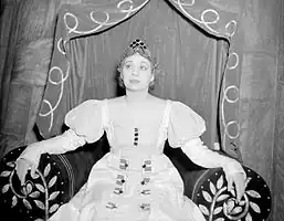 A woman wearing a crown and a white dress sits on a throne