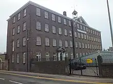 A four-storey brick building, on the long side to the right there are 17 windows on each floor, on the short side there are only 3 window each floor. Over the central five windows is a triangular pediment surmounted by a cupola with a bell. Over the ground floor door, four bays from the near corner is the date 1790.