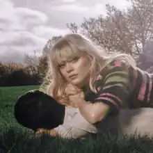 A woman with blonde hair lying on the back of a man with black hair, resting her arms on his shoulder-blades and looking towards the camera