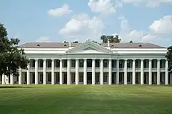 A wide white two-story building with many pillars. There is a portico for the length of the building with a second story balcony. There is a large lawn in front with some trees in back.