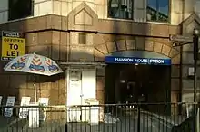 A beige-bricked building with a rectangular, dark blue sign reading "MANSION HOUSE STATION" in white letters and a yellow sign reading "OFFICES TO LET"