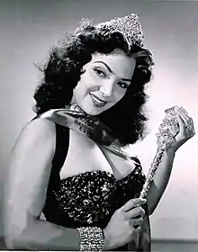 María Victoria smiling while wearing a black gown, a crown, and holding what it looks to be a sceptre