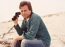 Photograph of Mark Lesser sitting on beach dunes, holding binoculars in one hand and smiling at the camera.