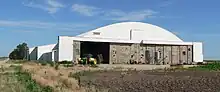 Large low building with plowed fields in foreground; two small grain bins in front of hangar; farmer working on tractor in hangar entrance