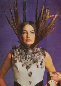 Woman wearing sleeveless top covered in small feathers. Long feathers attached to the collar stick up well past her face.