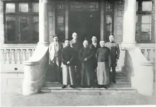 A black and white photograph of seven men, several wearing oriental clothing, standing on the entrance steps of a grand building.