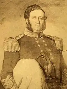 Sepia photo of a man in a fancy military uniform with heavy gold epaulettes and gold braid on cuffs and throat piece, holding a hat with ostrich plumes