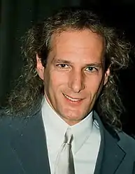 Middle-aged Caucasian man with long, blond hair, smiling while looking directly into the camera. He is wearing a collared shirt, suit and tie.