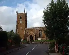 Red-brown church with limestone pinnacles and details. A square tower on the left, the nave on the right under a low roof hidden behind castellations in the same material. We can see a neat porch and a very imposing stained glass window. The church and graveyard stand a metre higher than the road, retained behind a low wall of the same red-brown ironstone. The church is seen framed by bushes and large trees in the middle nearground. The sky is mostly of high, light, clouds with patches of blue.