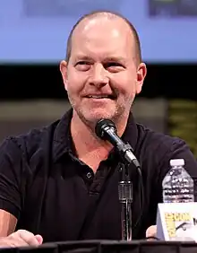 A man with close-shaven hair, and a slight beard, sits at a table before a microphone.