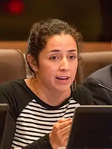 A dark-haired woman with double-hoop earrings and a black and white striped shirt speaks into a microphone from behind a computer screen.