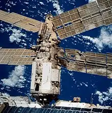 A gold-coloured solar array, bent and twisted out of shape and with several holes. The edge of a module can be seen to the right of the image, and Earth is visible in the background.