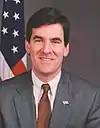 White male with dark hair in a gray suit with an American flag in the background