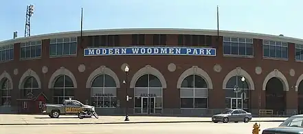 A large brick stadium with many round windows on the bottom and many rectangle windows in groups of four line the top of the stadium. The words Modern Woodmen Park are displayed above the door