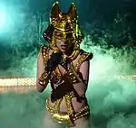 A woman in a shiny gold outfit, with golden gloves and a golden headress with two protrusions from the sides. Behind the woman, smoke can be seen billowing around.