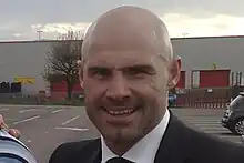 A man in a jacket and tie standing in a car park outside a football stadium.