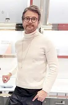 Image of designer Moritz Glik wearing a white cashmere sweater, a long necklace with a globe pendant, and reading glasses