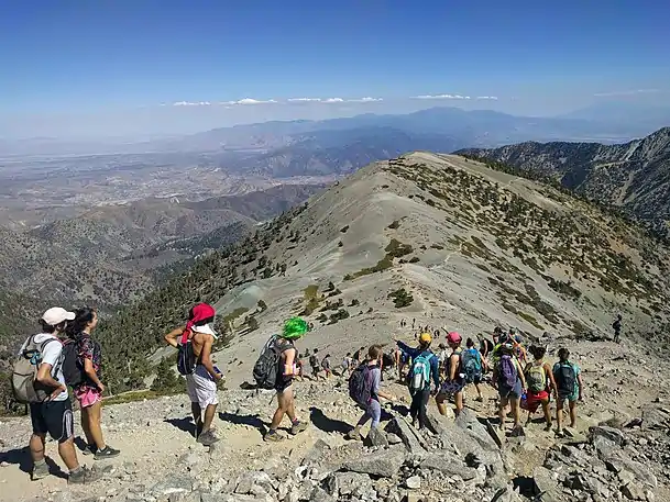 A line of students, many wearing costumes or swimwear, descends toward an alpine ridge