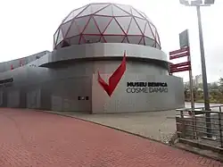 An outside view of the Benfica Museum. Its façade consists of grey metal panels with a red wing- or eagle-shaped piece of art followed by "MUSEU BENFICA COSME DAMIÃO" in black letters. On the top of the building, there is a red geodesic dome with a grey covering.