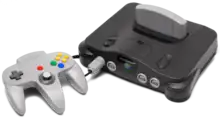 On the left, a three-pronged, handheld controller with a central analog stick and multiple buttons. On the right, a black electronics unit that accepts a light gray cartridge on the top and controllers (via ports) on its front.