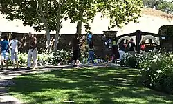 A group of people standing outside a gated area. There are trees, bushes, and grassed areas. A majority of the area the people and in are shadowed by the trees by the gate.