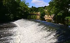 A weir on a wide river, with an arched bridge in the background