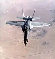 Jet fighter over-flying desert. The jet has black nose and two outward-canted vertical stabilizers between which lay two engines.