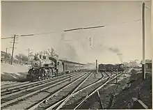 A view of a rail yard adjacent to mainline tracks. A steam locomotive pulls a passenger train on the main line heading towards the photographer, while in the yard tracks another locomotive is seen switching cars. Several other yard tracks are occupied with freight cars. Telltales are visible in the foreground above the tracks.