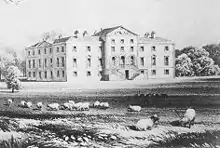 A house seen from an angle. The main front has nine bays, the central three of which protrude forward, and a double staircase leads to the main entrance on the first floor. Sheep graze in the foreground.