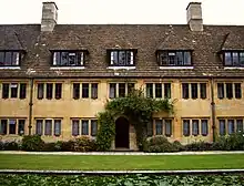 In the foreground, part of the long pool and lawn; behind, a section of a two-storey building in light stone, topped with a dark tiled roof and two chimneys; there are dormer windows in the roof; the windows at ground and first floor level are regular and frequent; an open archway surrounded by vegetation in the centre