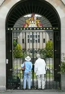 A large gate of metal bars, within a curved arch of light-coloured stone. Two people in casual clothes look through the gate, through which there is a grassed lawn with a pond, steps at the far end and a stone building with large rectangular windows. On the top of the gate, a shield with a coat of arms surmounted by a helmet.