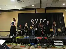 Photograph of several people on stage in front of a black curtain bearing the letters O V F F.