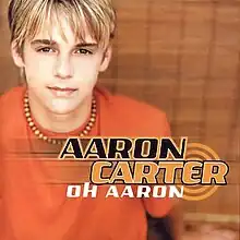 A young male adult with blonde hair is wearing an orange t-shirt and a bead necklace. The artist's name is colored in black and orange, and the album title is colored in white.