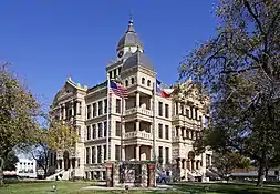 A stone building with a cloudy sky in the background. Three floors are shown with windows on each floor. There's a door entrance on the first floor and a large clock on the tower overhead.