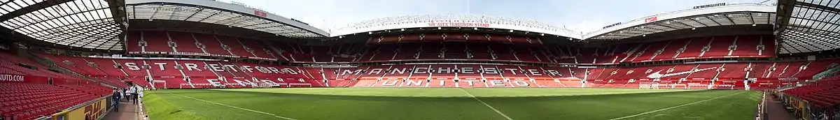 A stand of a football stadium. The seats are red, and the words "Manchester United" are written in white seats. The roof of the stand is supported by a cantilever structure. On the lip of the roof, it reads "Old Trafford Manchester".