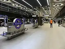 A wide island platform, with glass platform edge doors at either side. There are purple roundels labelled Paddington and benches in the centre. The ceiling is concrete with large circular light fixtures.