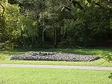 Elevated view of the cairn in the middle distance, from its side, with deciduous trees in leaf to its rear. To its front passes a wide asphalt path, dissecting flat ground of short grass. The tumulus' trapezium shape is evident, its boulders retained by a short wall, missing at the very front, left, where the rubble has tumbled out.