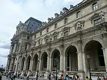 A classical building with ornamental design rises above a small crowd. Rounded archways line the front of the structure.