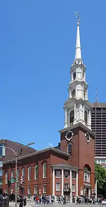 Ground-level view of a brick church with a large, white, tapering spire; a brown skyscraper is visible in the distance, with several shorter high-rises located closer to the church.