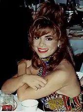 A woman sitting at a table with her arms crossed; she is wearing red lipstick and a colorful dress with sequins.