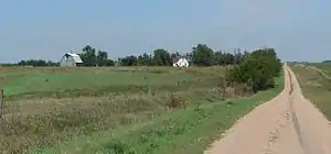 House and barn set in a line of trees in slightly rolling open country; gravel road runs by them