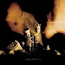 Two skeleton dolls wearing crowns stand in a darkened room with rocks. Orange glows are seen through the image.