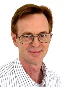 A man with glasses and white-and-black-striped color shirt stands in front of a white background.