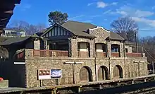 A stone building with a peaked roof and half-timbered pediments seen from its right, with a platform and some railroad tracks visible in the front. Three large round arched entryways at track level have been closed with wood paneling