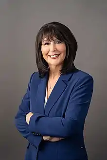 Philomena V. Mantella poses in front of a studio backdrop for a portrait. She stands, smiling, with her arms crossed, in a blue suit.